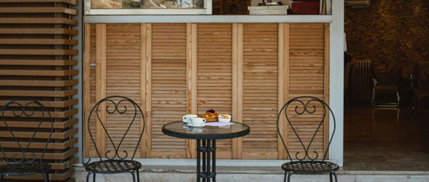 Cozy café setting with two wrought iron chairs and a small round table. The table holds a cup of coffee and pastries. Wooden panel backdrop adds warmth.